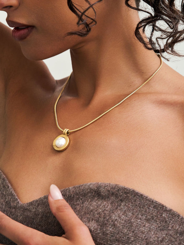 A close-up lifestyle shot of a model wearing a delicate gold chain necklace with a small oval pendant featuring a white pearl.