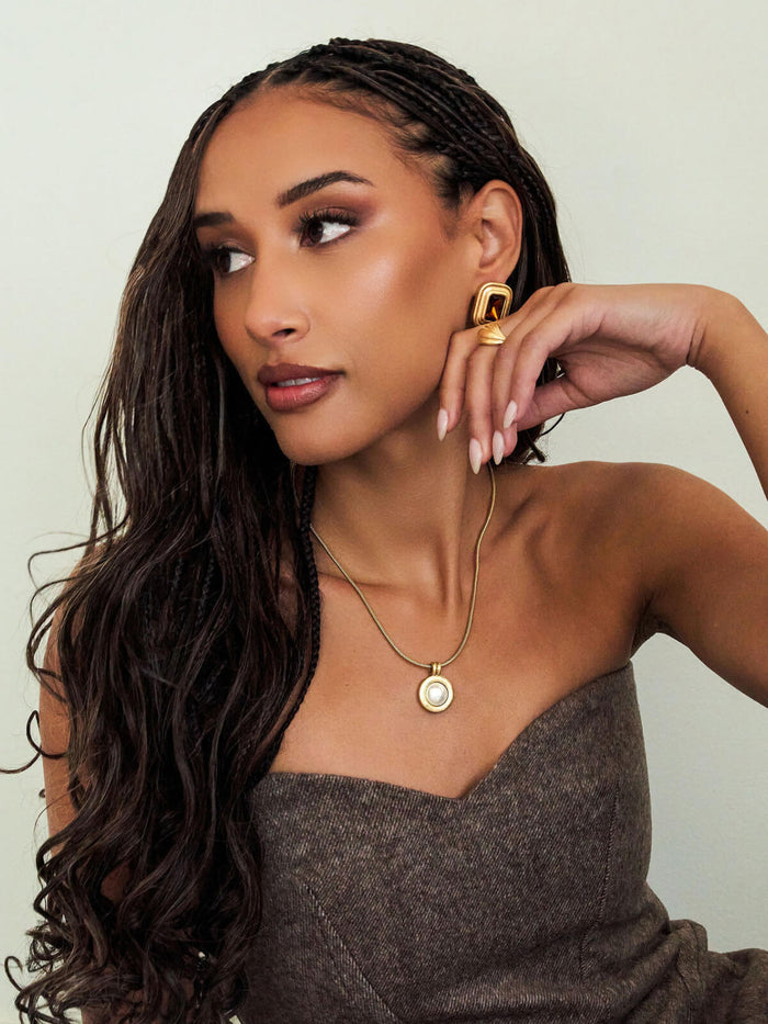 A woman with long dark curly hair models a gold pendant necklace with a white pearl-like center and a matching gold ring with a geometric gold bar. She is wearing a brown strapless top against a light beige background.
