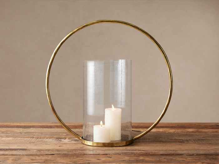 Circle Hurricane: gold circular frame holding a clear glass cloche with two lit white pillar candles on a rustic wood surface.