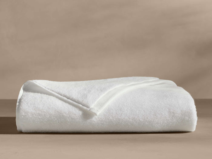 A neatly folded white towel with visible corner folds, lying on a beige surface, against a blurred beige background.