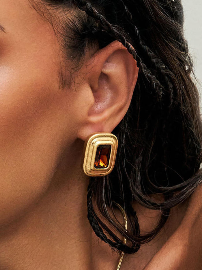 A close-up of a person's ear wearing a gold-toned rectangular stud earring with a faceted amber-orange center stone.