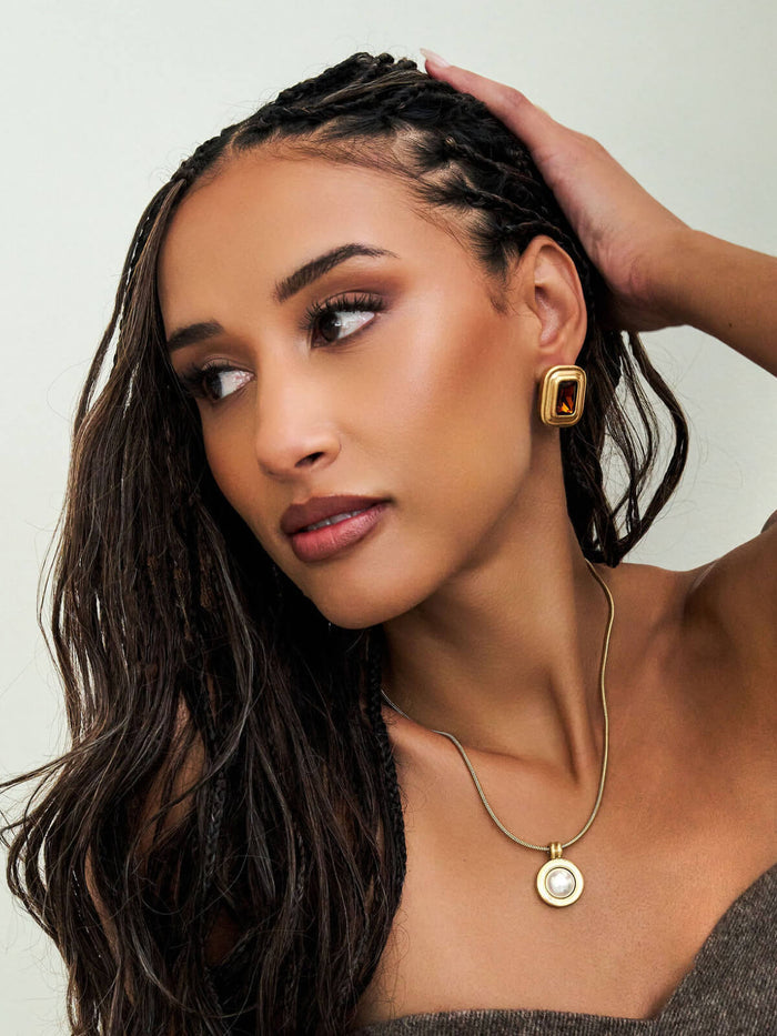 A close-up lifestyle shot of a woman with dark braided hair wearing a gold geometric drop earring and a gold pendant necklace with a white circular center, set against a plain light gray background.