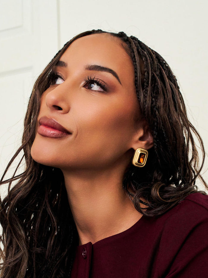 A close-up portrait of a woman with dark braided hair wearing a gold rectangular drop earring with a black center, against a light off-white background.
