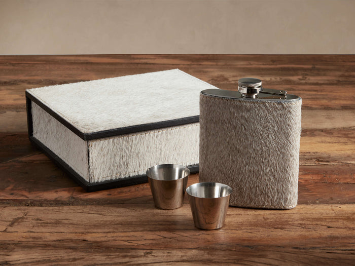 Colette Flask Set: A grey hair-on-hide flask, two silver shot glasses, and a white hair-on-hide storage box on wood.
