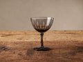 Cora smoky grey cocktail glass with etched vertical details on a rustic wooden table.