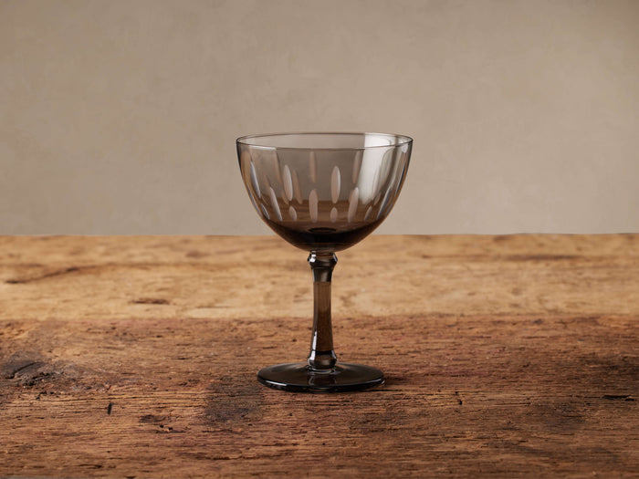 Cora smoky grey cocktail glass with etched vertical details on a rustic wooden table.