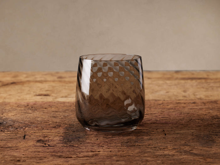 Cora Stemless Wine Glass, smoky grey with textured wavy and dimpled patterns, standing on a rustic wooden table.
