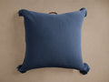 Square blue cotton knit floor pillow with visible texture, four tassels on corners, and a brown leather handle on top.