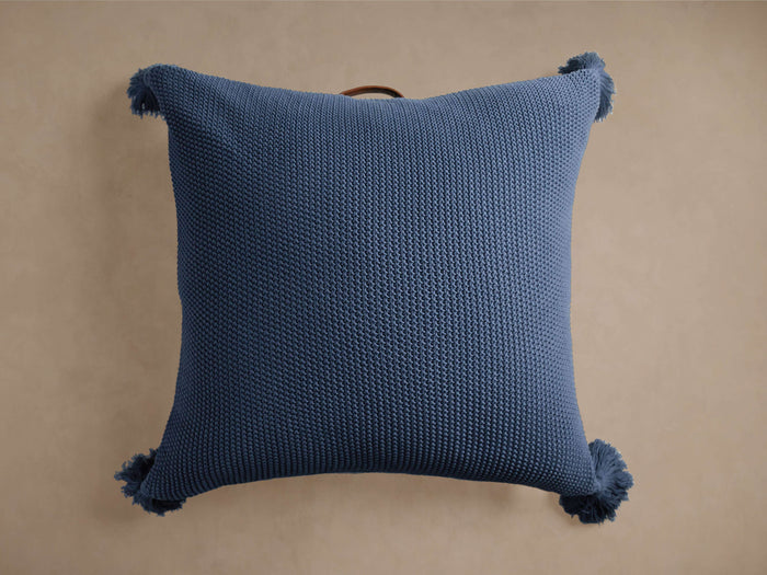 Square blue cotton knit floor pillow with visible texture, four tassels on corners, and a brown leather handle on top.