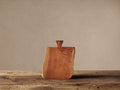Reclaimed wood cutting board, warm reddish-brown with natural grain, standing upright on a rustic wooden table. It has an irregular shape and a small triangular handle with a hole.