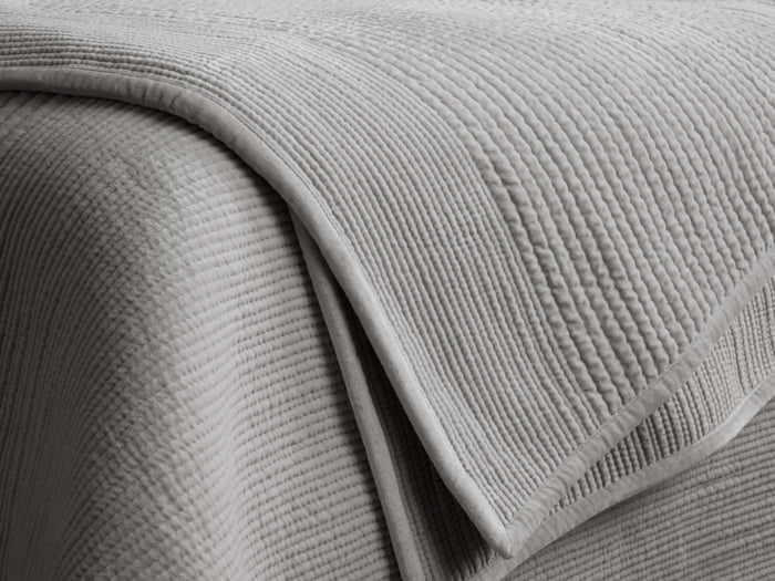 Daria Velvet Quilt, draped in light gray, showing its plush, horizontally quilted texture and soft corded piping.