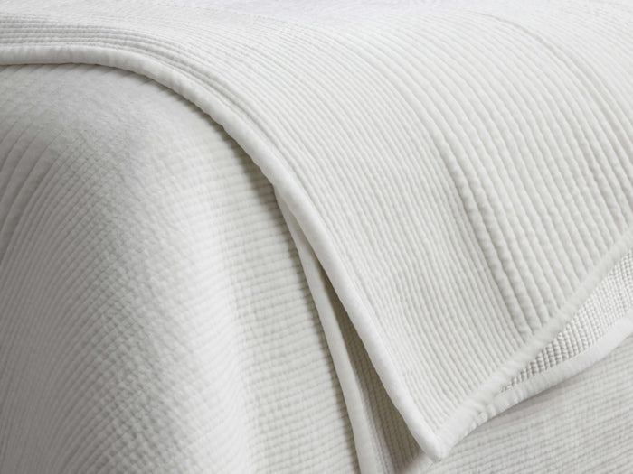 White Daria Velvet Quilt, plush and textured with parallel quilting lines. Folded over, showing corded piping trim.