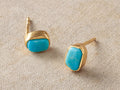 Two Darian Turquoise Bezel Stud Earrings: natural teal turquoise stones in organic gold settings, posts up, on light fabric.