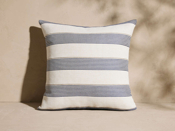 Dawson Outdoor Pillow, cream square with horizontal navy and tan woven stripes, upright against a beige textured wall.