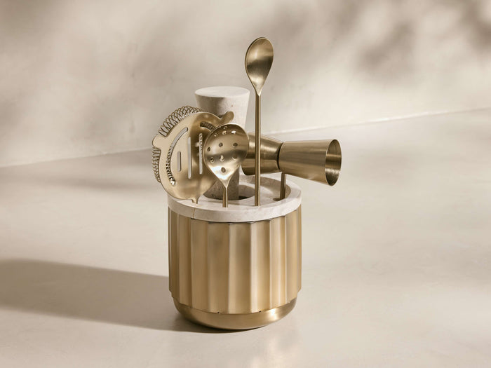 Front view of the golden Dayan Bar Tools set, including a fluted brass holder with a white marble top, strainer, jigger, and spoons.