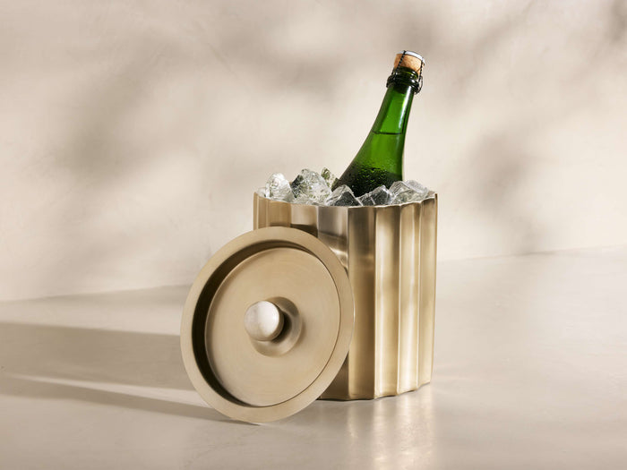 Fluted brass Dayana Ice Bucket holding a green champagne bottle with ice. Its lid, with a white marble handle, rests beside it.