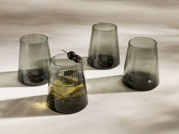 Four Della All-Purpose Glasses in smoke gray, reeded. One contains a golden drink with ice & a double olive skewer.