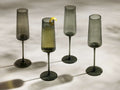 Four smoke-colored Della Champagne Flutes, reeded design, one with bubbly drink & lemon peel, arranged on a light surface.