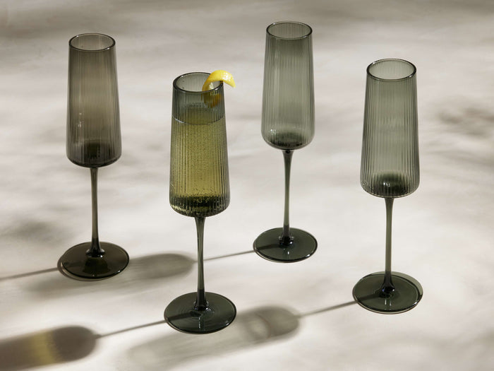 Four smoke-colored Della Champagne Flutes, reeded design, one with bubbly drink & lemon peel, arranged on a light surface.