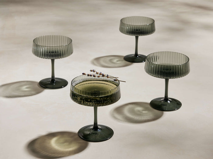 Four smoke reeded Della Coupe Glasses on a light surface. One holds a bubbly drink with a lavender sprig, casting textured shadows.