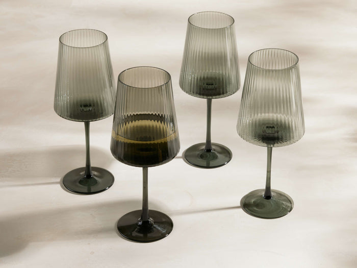 Four smoke Della wine glasses with reeded bowls, thin stems, and dark bases on a light background. One glass has liquid.