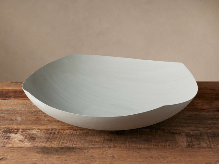 Delos Bowl, a large, wavy light gray decorative bowl, sits angled on a dark wood surface against a muted tan wall.