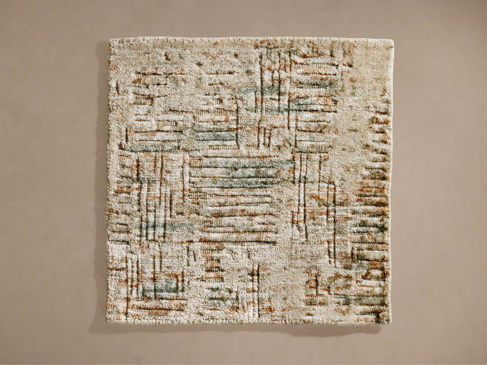 Devi Rug Swatch. Rectangular swatch with textured cream base, abstract brown and muted green lines, against a beige wall.