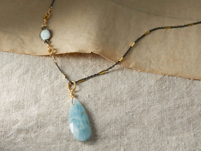 Devyn Necklace on fabric backdrop, features oxidized silver chain with gold accents, opal, and large aquamarine teardrop pendant.