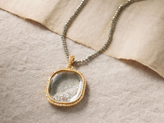 Diamond Shaker Cush Necklace features a gold-plated pendant with loose diamonds on a pyrite bead chain, lying on fabric.