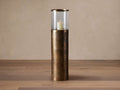 Dorian Small Candle Stand: Tall modern floor hurricane. Distressed antique brass textured base, clear glass cloche, lit white candle.