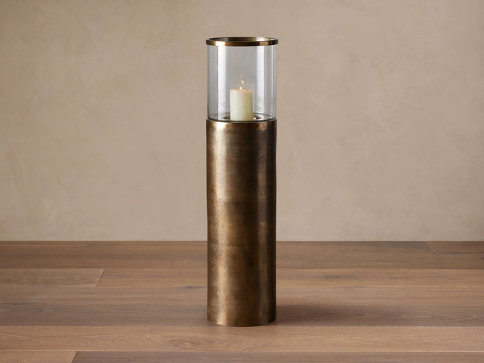 Dorian Small Candle Stand: Tall modern floor hurricane. Distressed antique brass textured base, clear glass cloche, lit white candle.