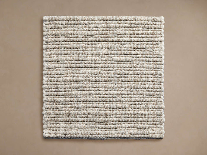 Dover Performance Rug Swatch, square, showing chunky, tightly woven light beige and brown textured fibers, flat on a tan surface.