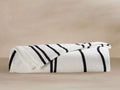 Folded Dunes Beach Towel, white with thick black vertical stripes in "Meteorite" color, lying on a light neutral surface.