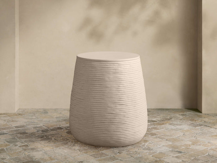 Eden Outdoor End Table. Light tan, round concrete table with distinct horizontal ribbed sides, on a stone patio.