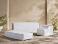 Tailored white Elio Outdoor Collection Covers on a patio sofa, chaise, and armchair. Beige wall with green plants in background.