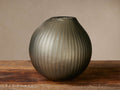 Elowen Vase: Round, smoky gray ribbed glass vase, artisan-crafted, on a rustic wooden table against a neutral wall.
