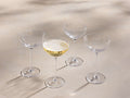 Front view of four clear Emrys Coupe Glasses, one filled with sparkling golden liquid, featuring geometric faceted bowls and slender stems.