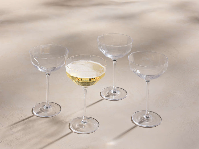 Front view of four clear Emrys Coupe Glasses, one filled with sparkling golden liquid, featuring geometric faceted bowls and slender stems.