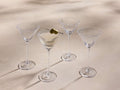 Front view of four clear Emrys Martini Glasses, featuring geometric lines. One glass holds a martini with two green olives on a pick.
