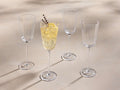 Set of four clear Emrys Wine Glasses with faceted bowls. One glass holds a yellow drink, ice, a lemon slice, and lavender sprigs.