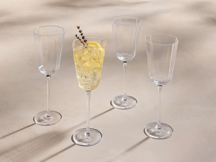 Set of four clear Emrys Wine Glasses with faceted bowls. One glass holds a yellow drink, ice, a lemon slice, and lavender sprigs.