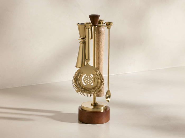 Front view of gold-toned Errol Bar Tools on a dark wood stand, featuring a jigger, strainer, and spoon.