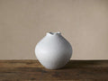 White, round Eva vase with textured surface and ruffled rim, sitting on a dark wooden surface against a light wall.