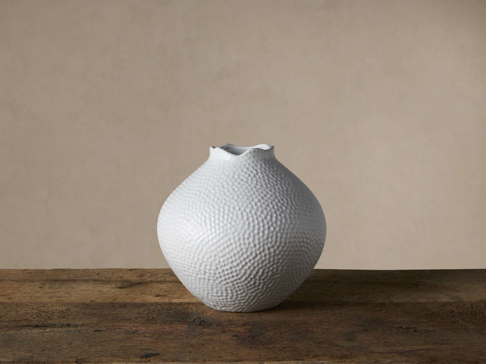 White, round Eva vase with textured surface and ruffled rim, sitting on a dark wooden surface against a light wall.