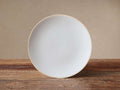 Round Evora Salad Plate, white glazed stoneware with an unglazed light brown rim, standing vertically on a rustic wood table.