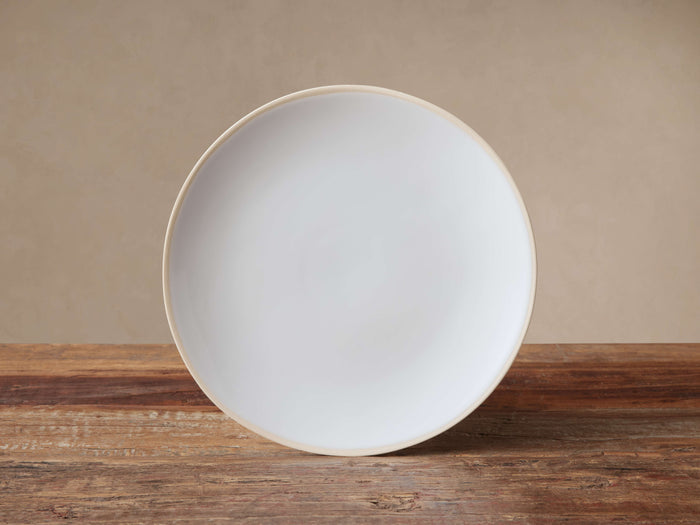 Round Evora Salad Plate, white glazed stoneware with an unglazed light brown rim, standing vertically on a rustic wood table.