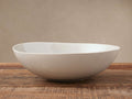 Evora Serving Bowl, an off-white stoneware bowl with a subtly curved rim, rests on a rustic wooden table against a beige wall.