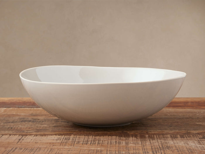 Evora Serving Bowl, an off-white stoneware bowl with a subtly curved rim, rests on a rustic wooden table against a beige wall.