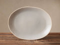 Evora Serving Tray: Oval, handcrafted light gray glazed stoneware, unglazed rim detail, angled on a rustic wood surface.
