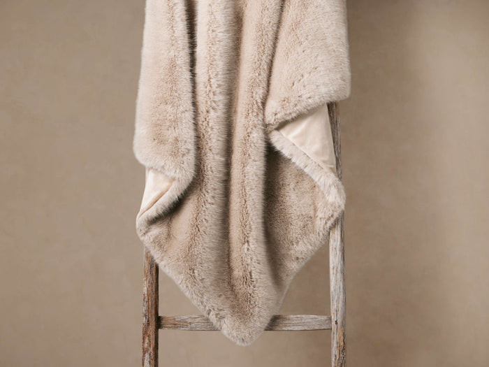Soft Fairbanks Faux Fur Throw in Dune, a light beige blanket, draped over a wooden ladder, showing plush texture and microfiber lining.